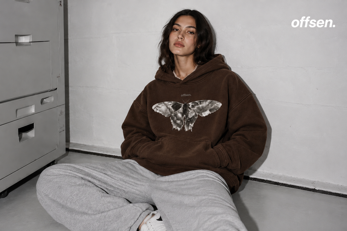 Model sitting on the floor against a grey wall in a brown OFFSEN hoodie with a large moth print and grey sweatpants